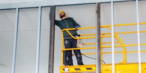 Construction worker wearing safety helmet and work gloves, standing on a mobile elevating work platform installing drywall on a metal frame